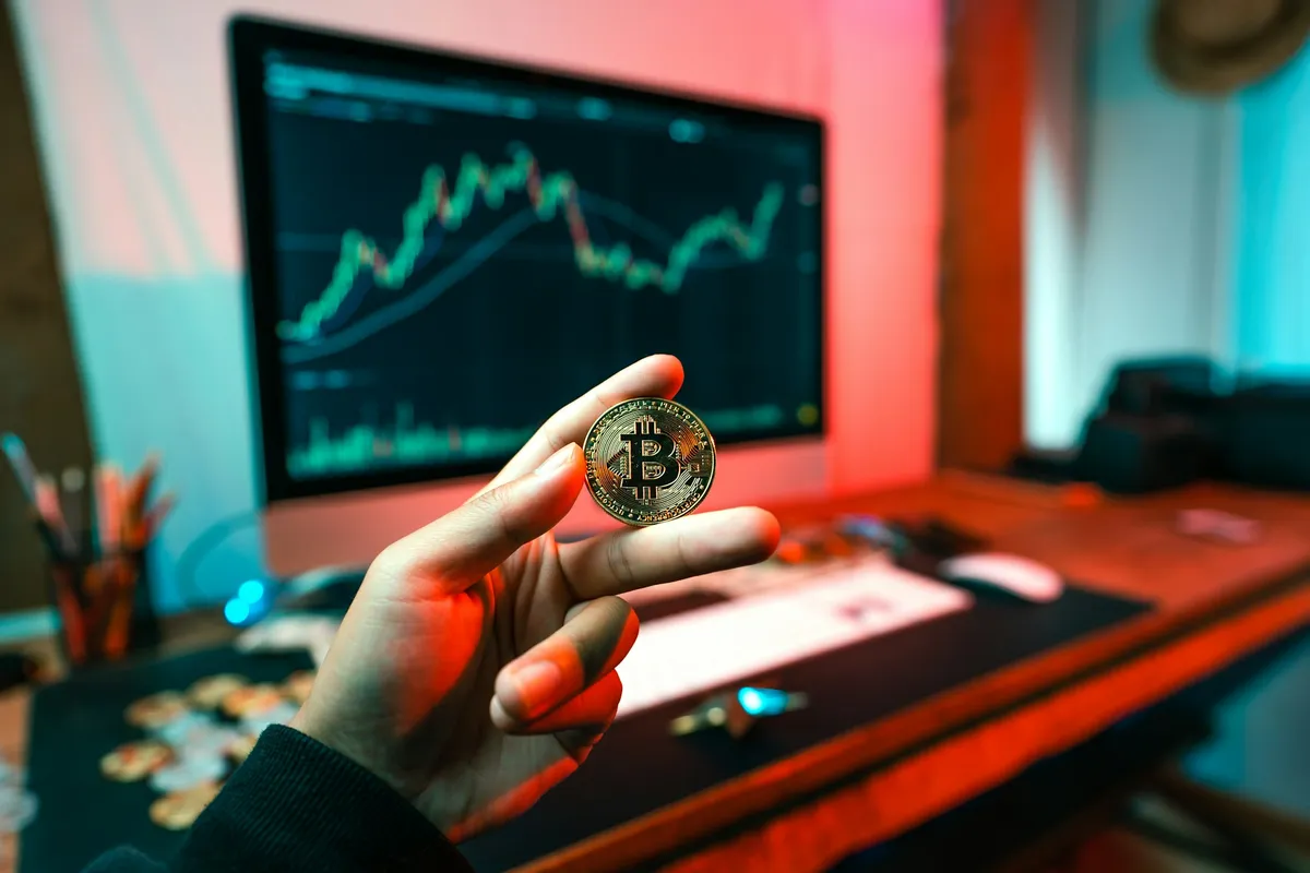 A person holding a Bitcoin coin in front of a computer